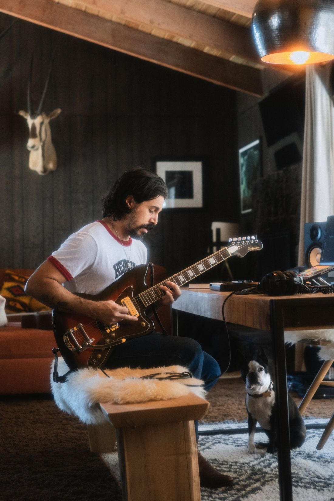 Coquí playing a sunburst electric guitar in a wood-paneled home studio while a Boston terrier looks on