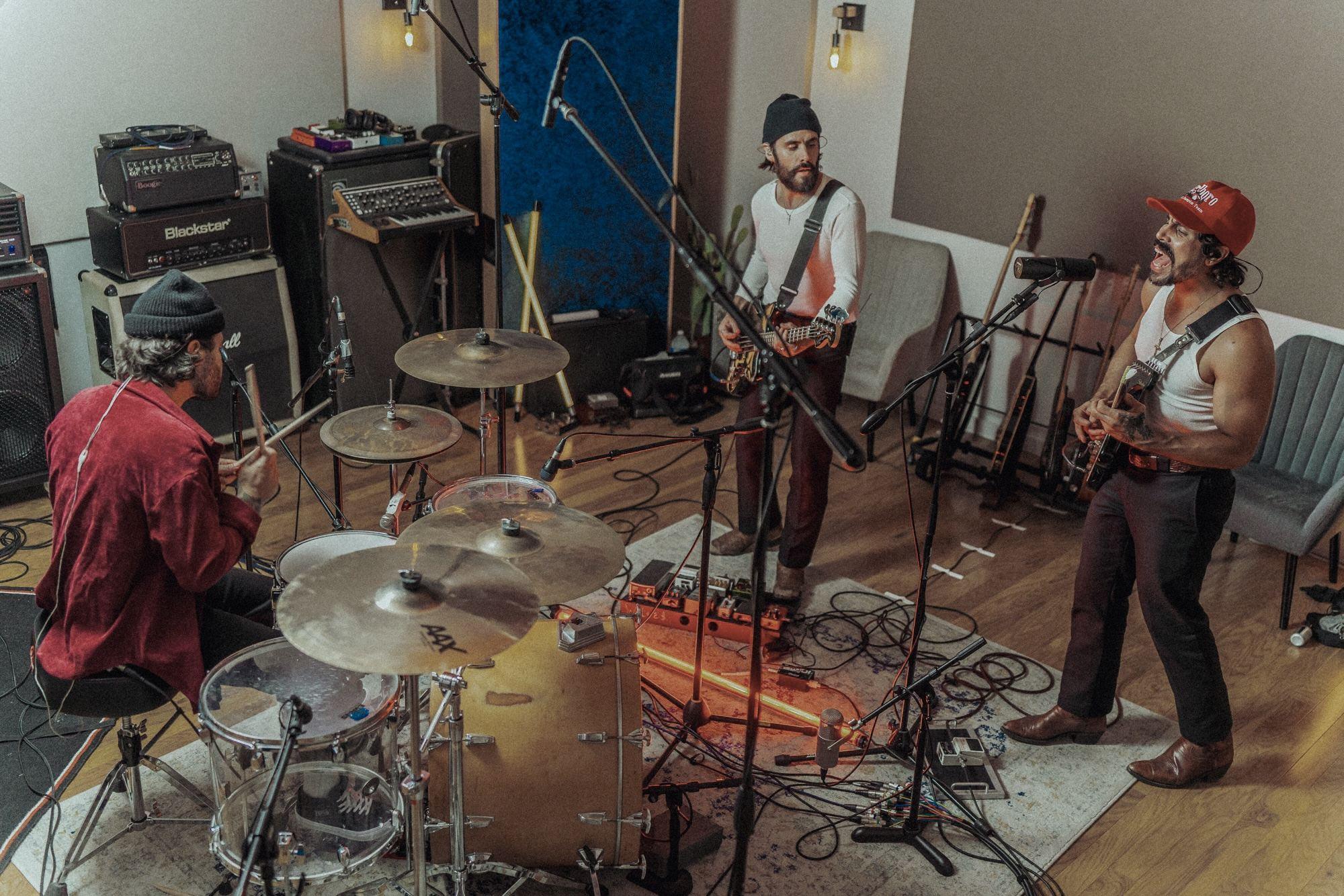 Three musicians playing in a studio, seen from above: a drummer on the left, a bass player in a beanie in the middle and a singer with a guitar in a red cap on the right